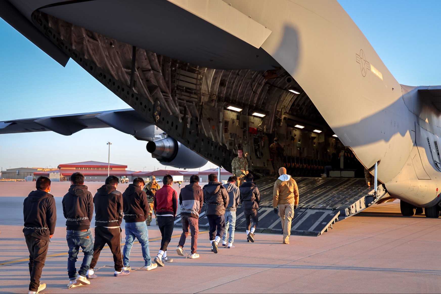 Personas migrantes siendo deportadas de los Estados Unidos. Dos filas de hombres esposados de las manos y encadenados de las extremidadees caminando para abordar un avión militar.
